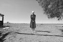 A woman in Amish dress and bonnet standing in a field looking down at a bottle and a pack of cigarettes, black and white photograph, Salton Sea, 1 May 2016